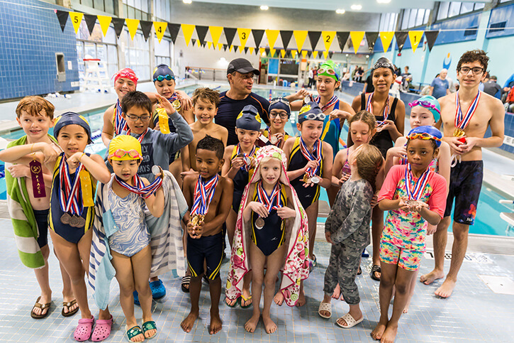 a large group of children in swimsuits standing next to a pool wearing competition medals around their neck