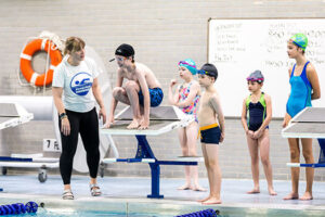 Instructor guiding a child on a starting block while other young swimmers wait poolside during a lesson