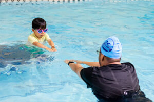 Child wearing pink heart-shaped goggles facing instructor in pool, practicing streamlining.