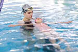 Instructor supports a child in pink swim gear floating calmly on their back in a lane of an indoor pool.