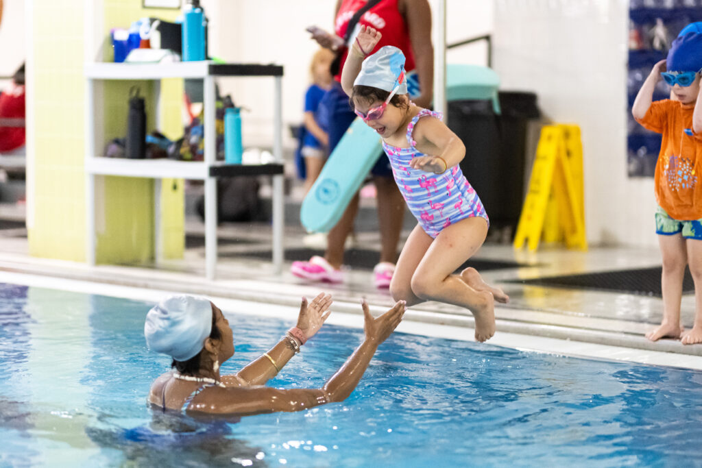 Young child in swimsuit and swim cap jumping into pool toward instructor ready to catch them during a swim lesson