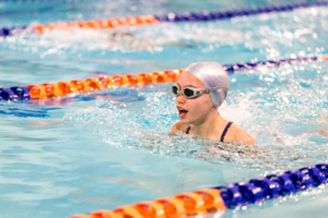 Swimmer in a blue indoor pool wearing a silver swim cap and goggles, racing in orange lane markers.