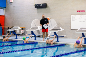 Swim coach standing on starting blocks instructing young swimmers in a pool practice, with a whiteboard and 'No Smoking' sign in the background.
