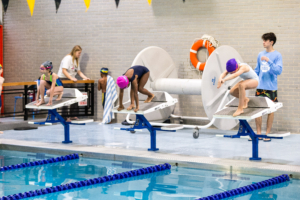 Swimmers on starting blocks preparing to dive in during a swim practice at an indoor pool.